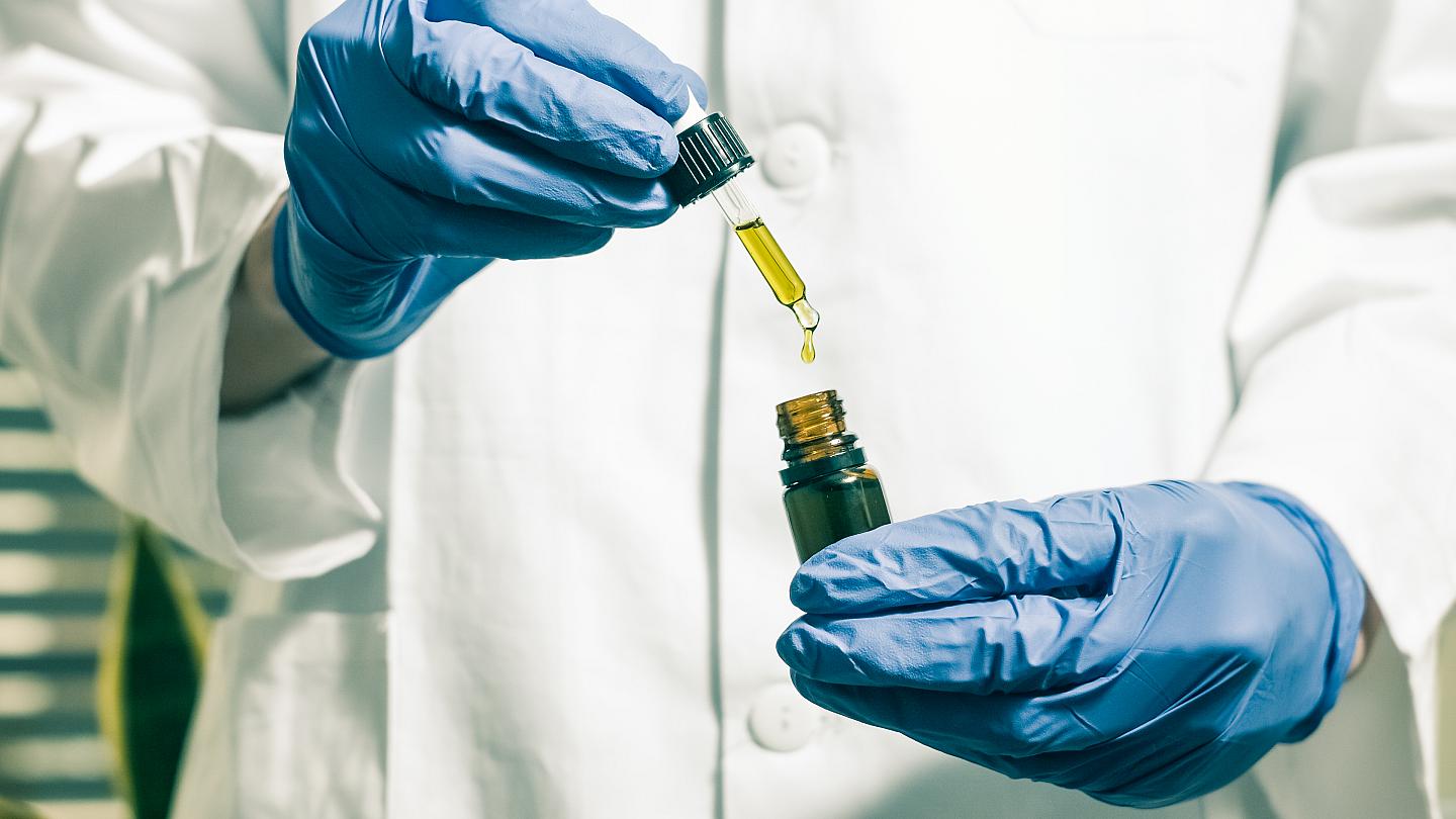 A man wearing a white lab coat with latex gloves in both hands squeezing a tincture dropper filled with CBD oil into a brown tincture bottle