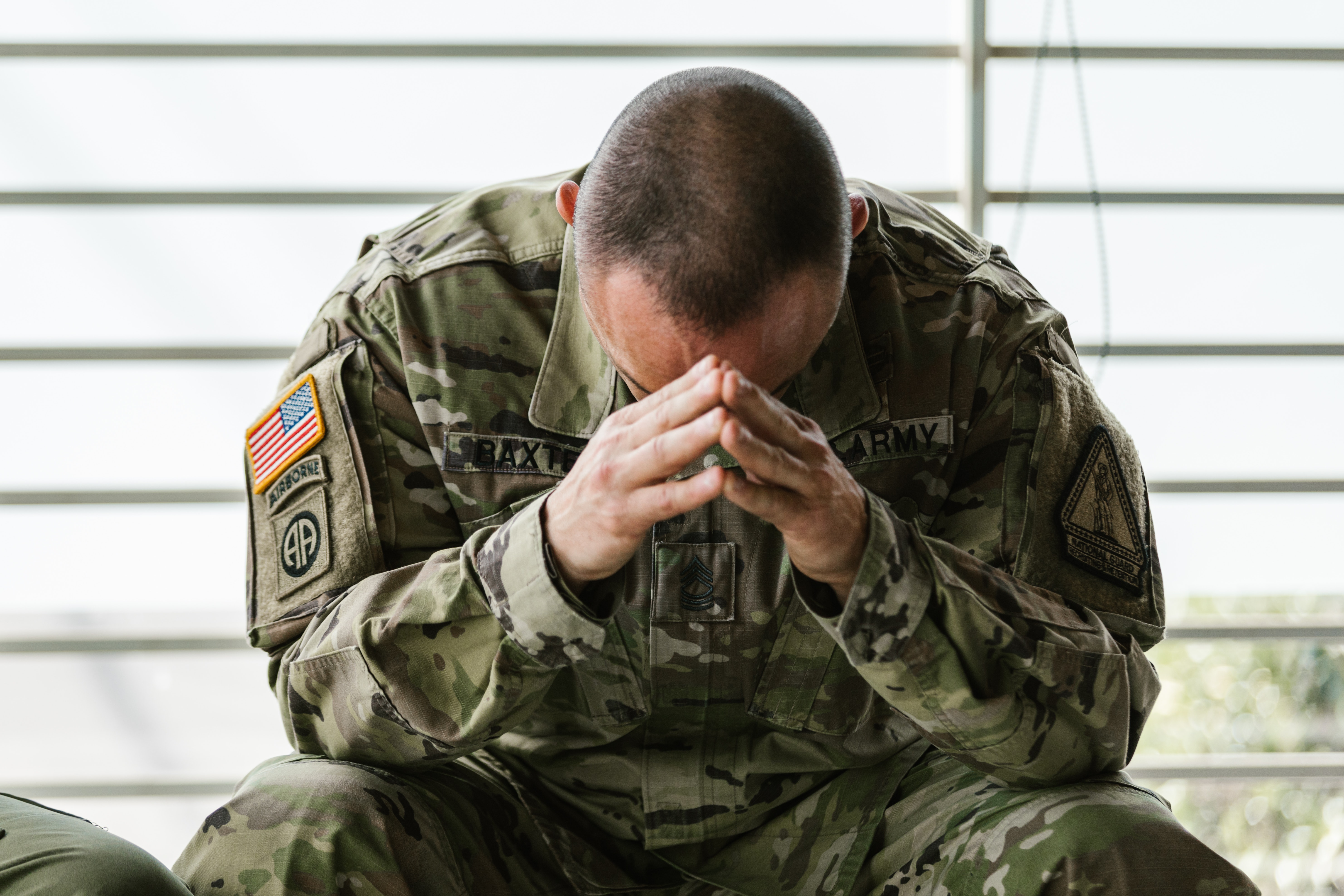 A Caucasian man wearing a military camo uniform sitting with both hands to his forehead while looking down