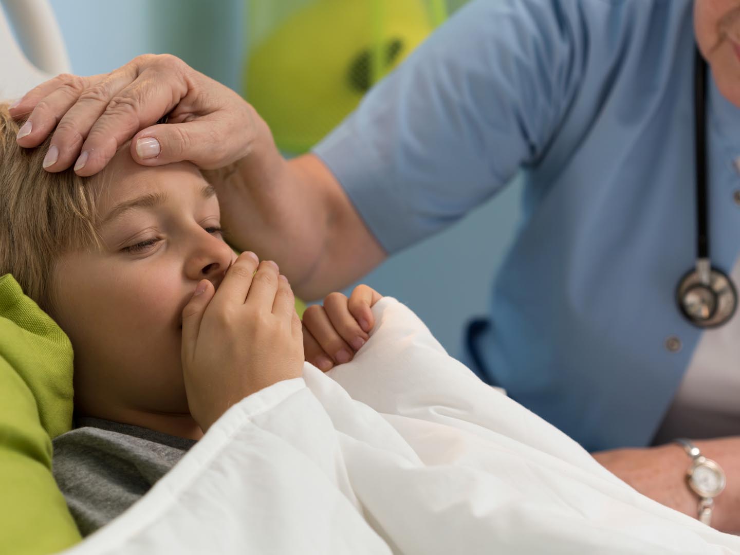 A girl laying down coughing with one of her hand covering her mouth and a nurse beside her with one of her hand on the girl's forehead