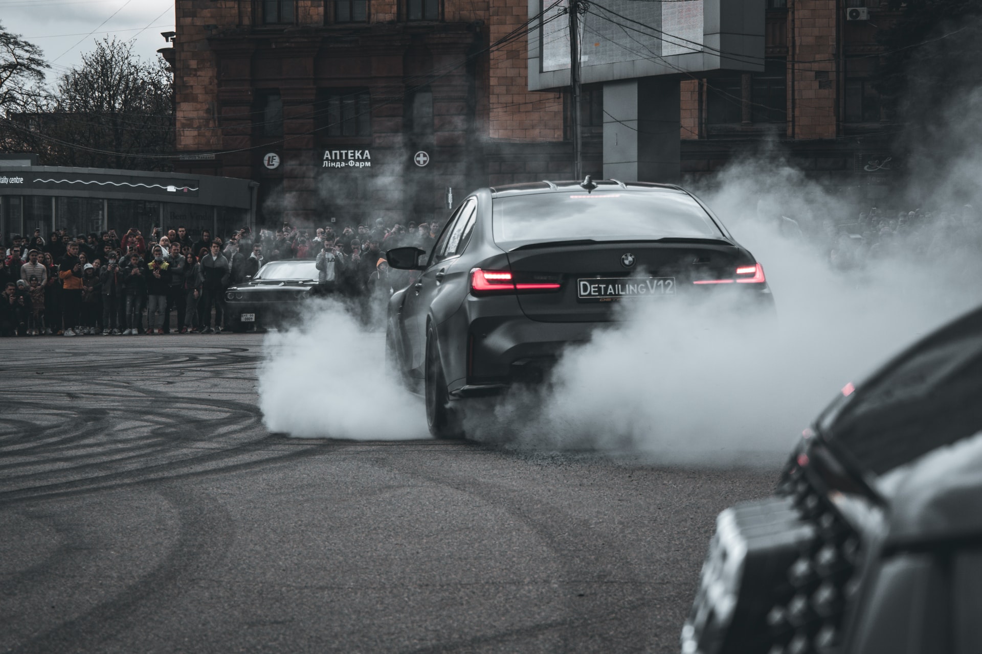A black car emitting smoke with a vehicle behind and people watching 