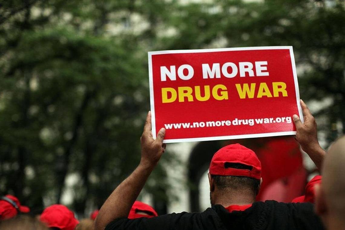 A man wearing a red face cap and a black t-shirt holding a placard with the word "No more drug war" above his head