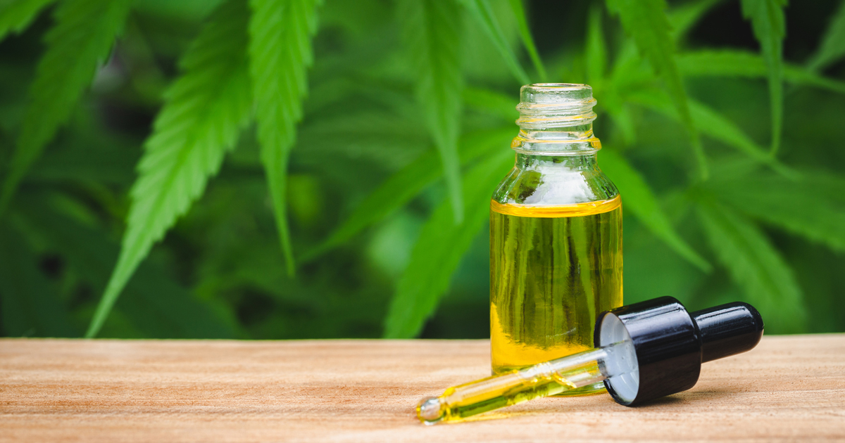 An opened transparent tincture bottle filled with CBD oil sitting beside its dropper on a wooden platform with cannabis leaves as the background
