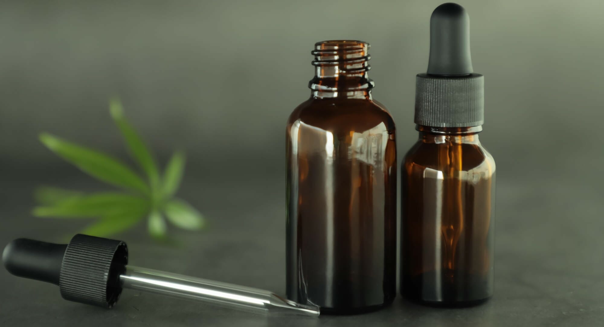 An opened brown tincture bottle with its dropper laying beside it sitting beside a closed brown ticture bottle with a blurred cannabis leaf in the background