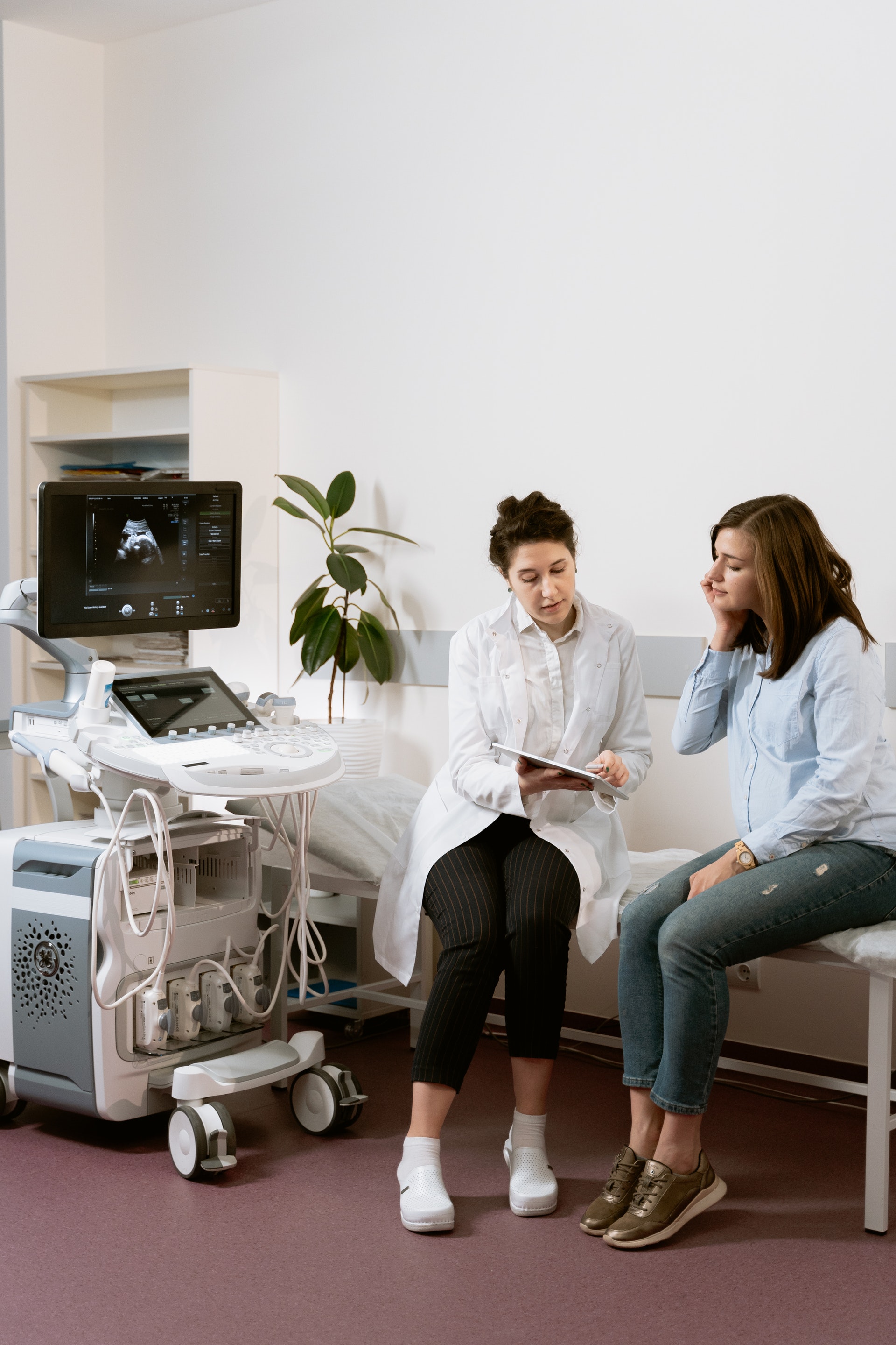 A woman doctor is talking to a woman on a patient's bed, and an ultrasound machine is present beside them