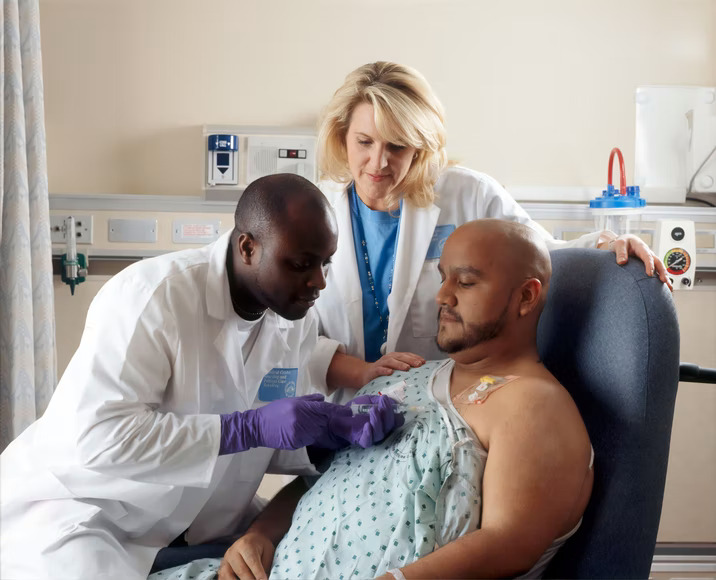 A Hispanic male patient received treatment from an African-American Nurse through a port that is placed in his chest area. A caucasian female nurse looks on