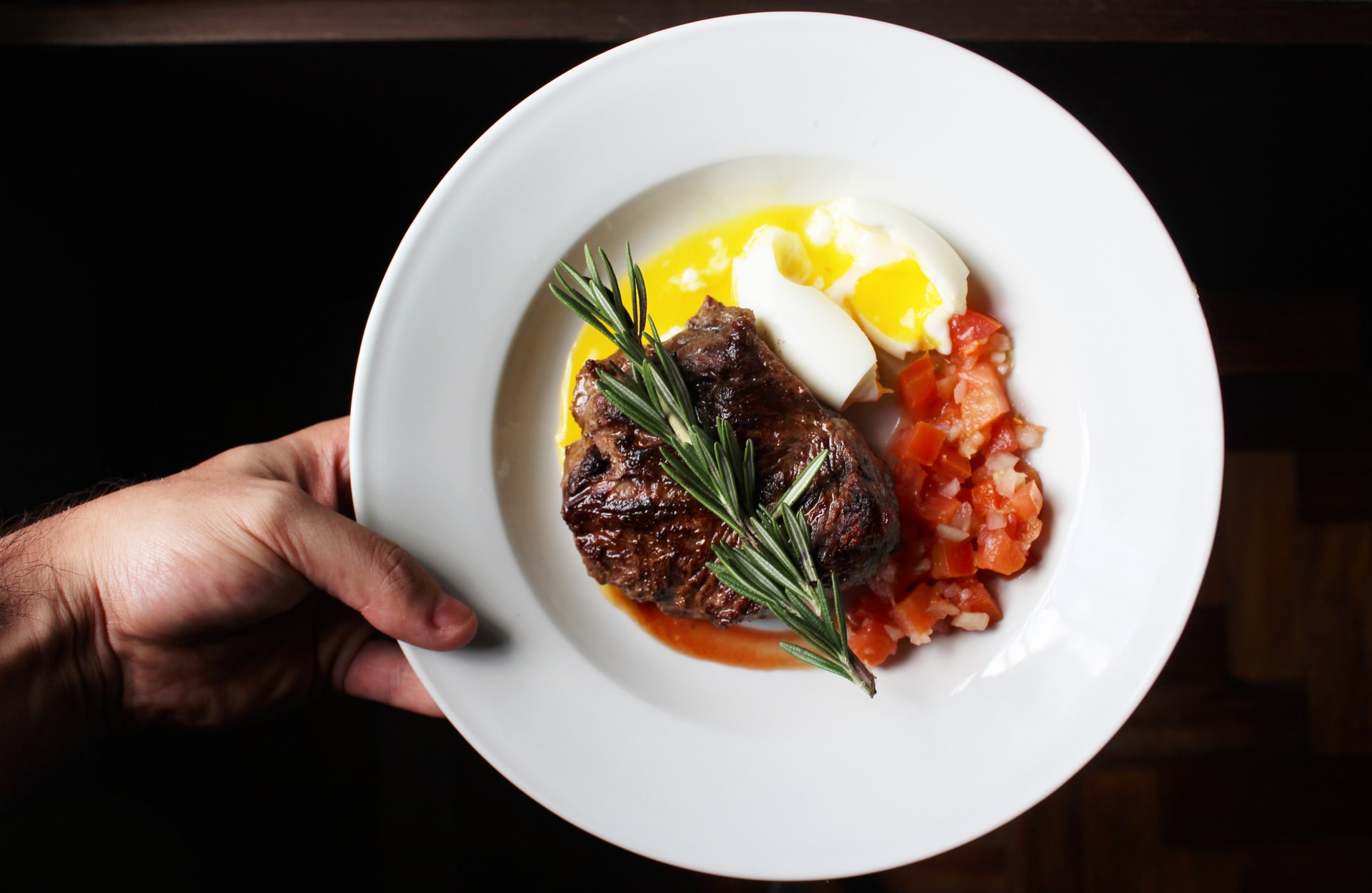A white hand holding a white flat plate filled with meat, some eggs, and vegetables
