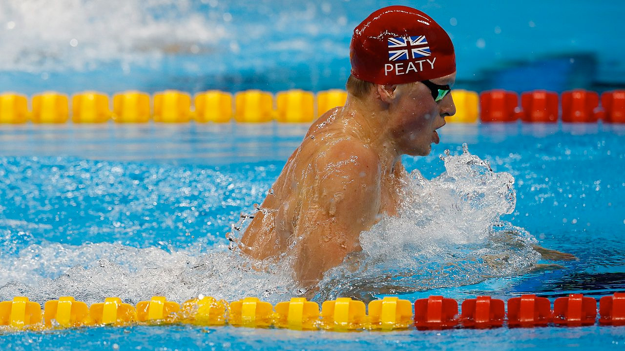 A male swimmer with a red swimming cap and glasses doing the breaststroke in a swimming pool