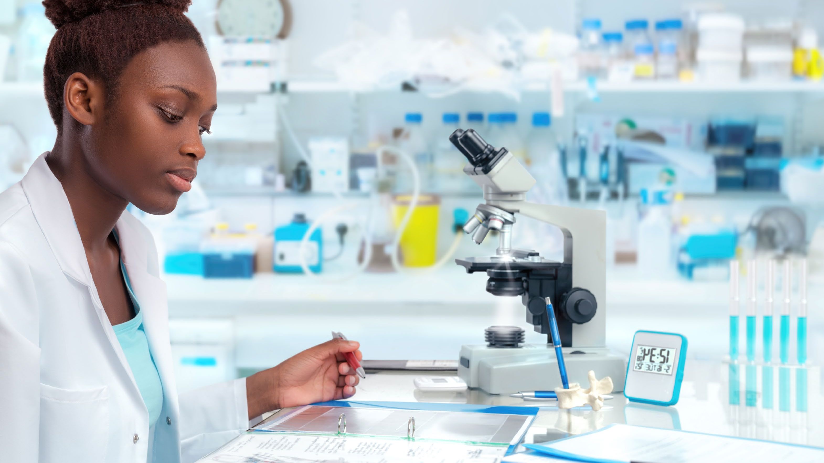 A black woman wearing a lab coat sitting at a table with a microscope on it with other lab equipment while holding a pen on her left hand and looking at her note pad