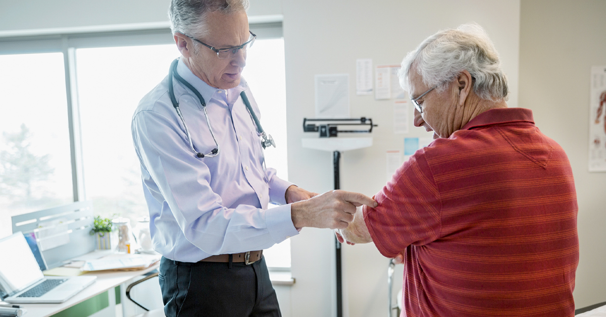 A doctor with a stethoscope round his neck doing a checkup on the arm of an elderly man wearing a red t-shirt