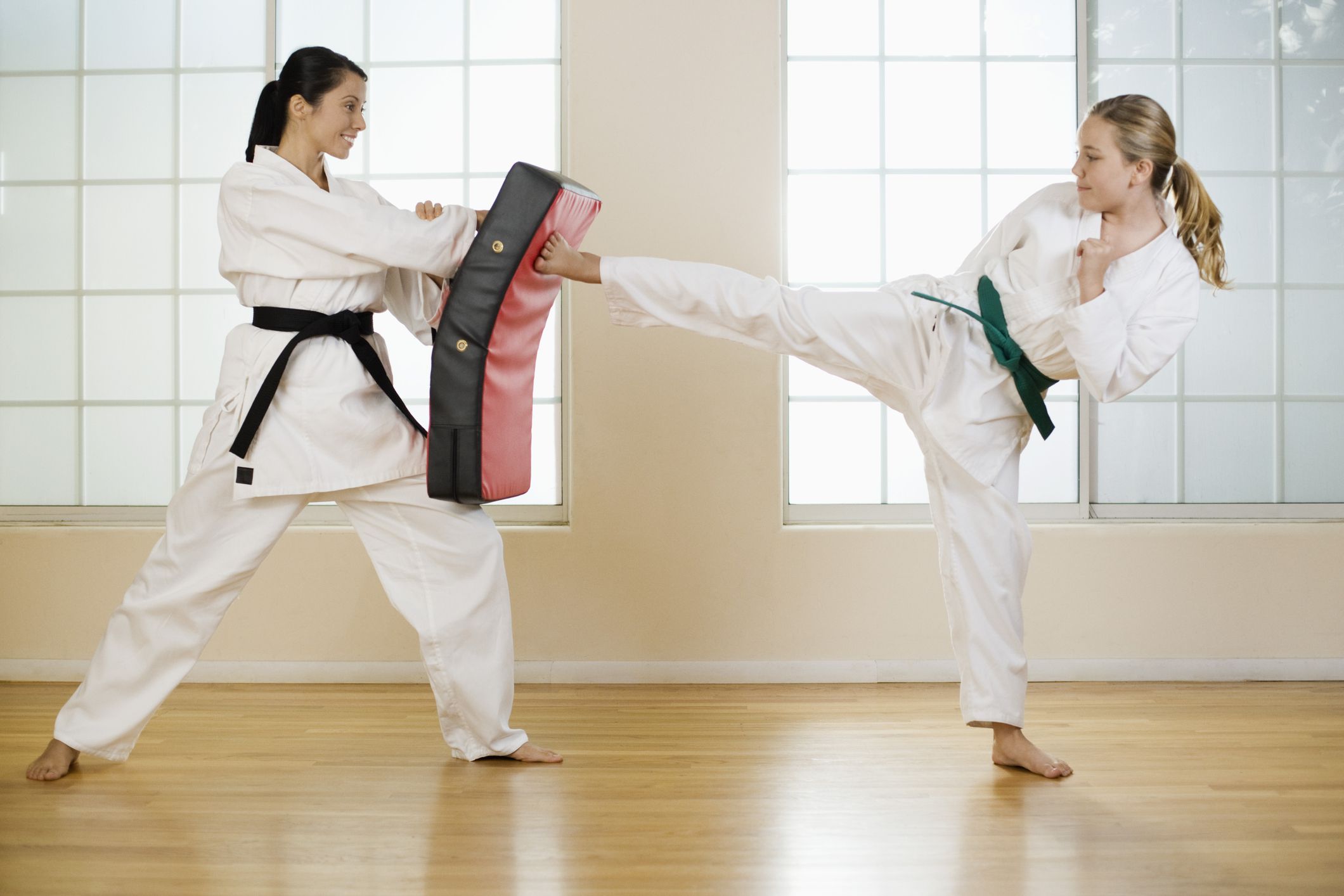 A woman with a green belt in karate kicking a bag while another woman with a black belt holds it
