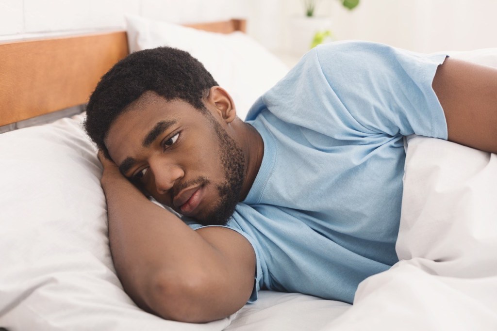A black man wearing a blue t-shirt laying awake on his bed while being covered with a white duvet