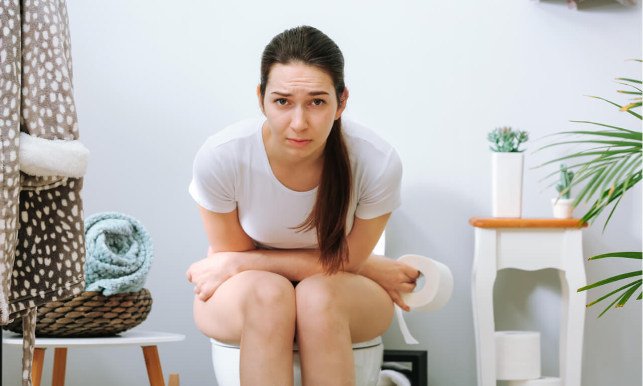 A woman seated on a toilet seat holding her stomach with a roll of toilet paper in one hand